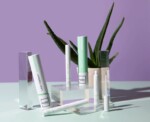 Several cosmetic products labeled "Lash Therapy" and "Hello Lashes" are arranged on clear stands with an aloe vera plant in the background on a pastel green surface.