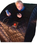 A woman smiles while using a portable infrared sauna. She holds a remote control in one hand and a smartphone in the other, peeking out from the top opening of the quilted black sauna.