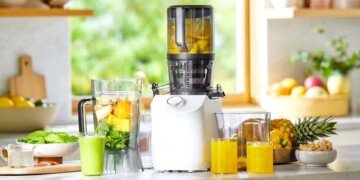 A modern juicer on a kitchen counter surrounded by whole and sliced fruits, with fresh juice in glasses nearby.
