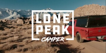 A red truck, with "Lone Peak Camper" text overlay, drives on a dirt road through a rocky desert landscape with mountains in the distance.