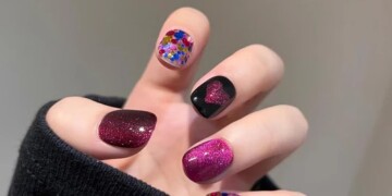 A hand displaying nails with colorful and glitter designs, including one with a black polish featuring a heart shape and one with a gradient effect. The word "TAGLESSNAILS" is visible above.